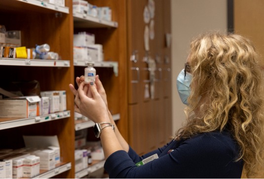 Shannon_Medicine-092821 1 (2) A woman wearing a medical mask holds up a vial of medicine in a storeroom